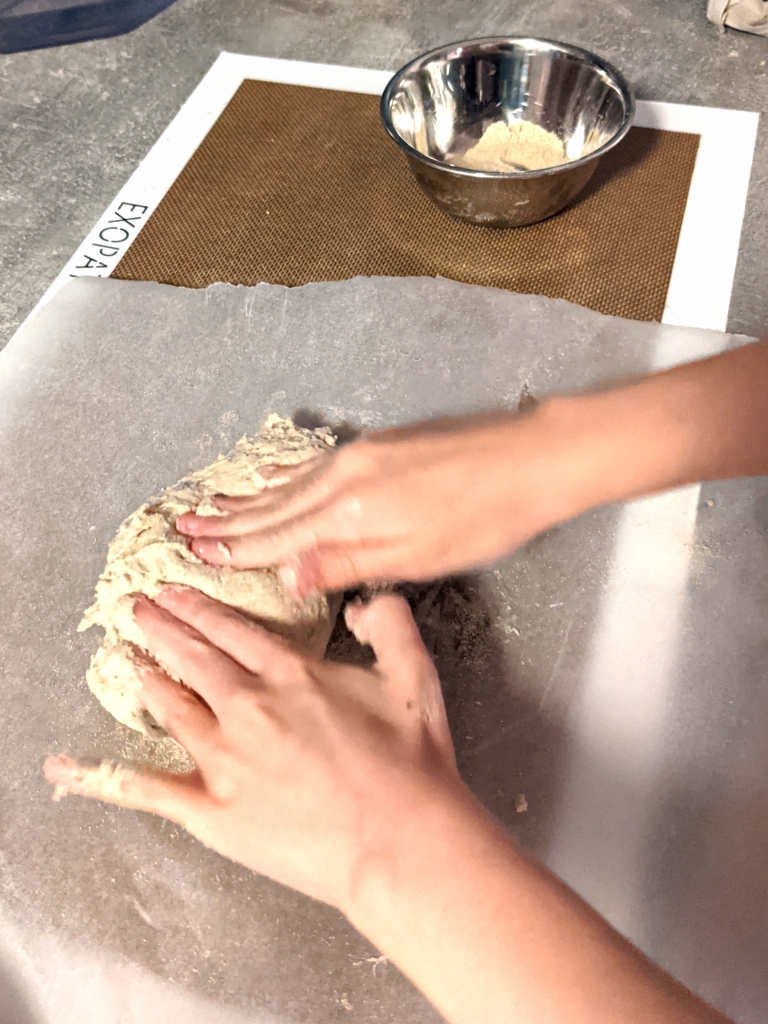 A close-up of a girl's hands kneading a piece of dough on top of parchment paper. There is some dough stuck in the fingers of the girl. The hands in the picture are a bit blurred showing motion.