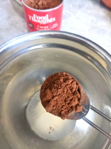 A tablespoon of dark cocoa powder above a silver bowl, cocoa tub in the background.